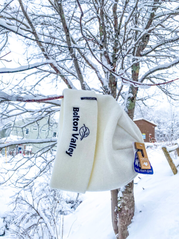 White embroidered beanie that reads "Bolton Valley" hanging from a snow covered tree