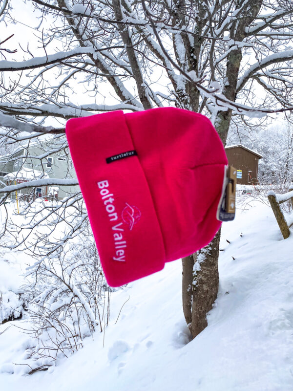 Pink embroidered beanie that reads "Bolton Valley" hanging from a snow covered tree
