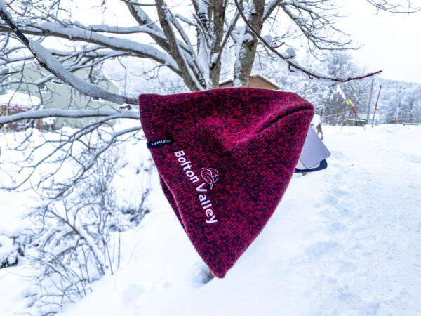 Red embroidered beanie that reads "Bolton Valley" hanging from a snow covered tree